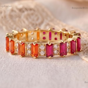 May include: A gold eternity band featuring alternating baguette-cut orange and pink gemstones, interspersed with small round clear stones. The ring displays a vibrant gradient of warm colors set in a continuous metal band design.
