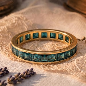 Puede incluir: Un anillo de oro con piedras preciosas cuadradas de color verde azulado engastadas alrededor de la banda. El anillo se exhibe sobre una superficie de encaje color crema, con lavanda seca y un libro antiguo en el fondo. El diseño del anillo es simétrico.