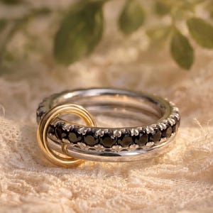May include: Three interlinked rings, two silver and one gold. The silver rings are set with black gemstones. The gold ring is a plain band. The rings are presented on a textured, off-white surface.