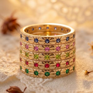 May include: A stack of gold-colored rings, each adorned with a repeating pattern of small, colorful gemstones. The gemstones include blue, pink, green, red, and emerald green. The rings are stacked on a white lace surface.