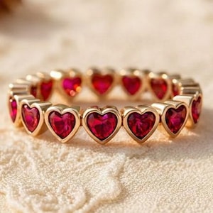 May include: A gold ring adorned with a sequence of heart-shaped, red gemstones. The ring is set against a soft, textured backdrop with dried floral elements, creating a romantic and elegant aesthetic. The ring is a piece of jewellery.