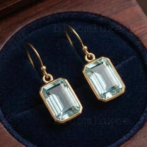 Può includere: Un paio di orecchini pendenti color oro con pietre preziose rettangolari azzurro chiaro. Gli orecchini sono esposti in un portagioie di legno scuro con interno in velluto blu navy. La scatola è aperta.