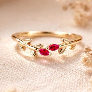 Op de afbeelding: Een goudkleurige ring met een bladeren ontwerp. De ring heeft twee ovale, helderrode edelstenen. De ring is geplaatst tegen een zachte, neutrale achtergrond met delicate bloemaccenten, wat een romantische uitstraling creëert.