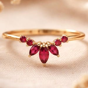 Könnte beinhalten: Ein goldener Ring mit einer fächerförmigen Anordnung roter Edelsteine. Der Ring hat marquise- und rund geschliffene Steine. Der Ring wird auf einem neutralen Hintergrund präsentiert. Die Edelsteine sind tiefrot.