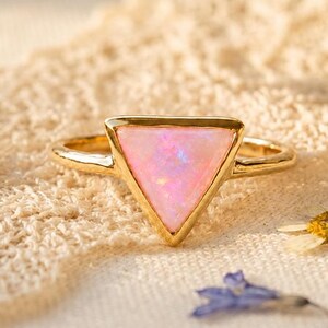 May include: A gold ring featuring a triangular-shaped pink opal gemstone. The stone displays iridescent flashes of light across its surface. The band is thin and polished, holding the triangular setting securely in place on the finger.