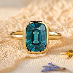 May include: A gold ring featuring a large, rectangular, teal-colored gemstone set in a smooth, polished bezel setting. The band is thin and circular, crafted from a warm-toned yellow metal that complements the deep blue-green hue of the stone.
