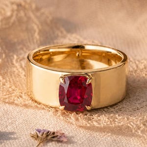 May include: A wide gold band ring featuring a single, cushion-cut, deep red gemstone set in a four-prong mounting. The polished metal surface of the ring reflects light, highlighting the vibrant color of the central stone and the smooth band.