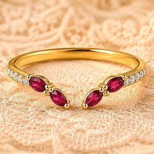 May include: A gold open-style ring featuring two marquise-cut red rubies at each end, accented by small clear round stones set along the band. The ring has a polished gold finish and a gap between the two ruby-adorned ends of the band.