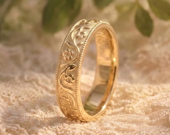 May include: A gold ring with an intricate floral pattern. The ring's surface is adorned with detailed carvings of flowers and vines. The ring is set against a soft, textured background, highlighting its elegant design. The ring is a symbol of commitment and love.