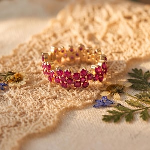 May include: A gold-colored ring with a floral design, featuring numerous small, round, ruby-colored gemstones. The ring is set against a textured, beige fabric, with dried flowers and greenery in the background.
