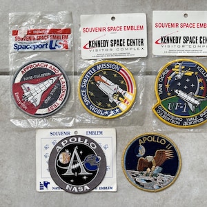 May include: A collection of six embroidered patches featuring NASA space shuttle and Apollo mission imagery. The patches are yellow, red, blue, and black with white lettering. The patches are labeled "Spaceport USA", "Kennedy Space Center Visitor Complex", "Approach and Landing Test", "100th Space Shuttle Mission", "Tani Gorrie Kelly Godwin UF-1", and "Apollo".