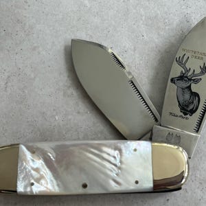 May include: A whitetail deer pocket knife with a mother of pearl handle and gold accents. The blade features an engraved image of a whitetail deer with the words "Whitetail Deer" and "Toad River" on the blade.