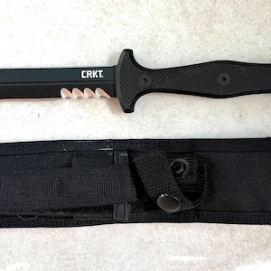 May include: A black CRKT tactical knife with a serrated edge and a textured handle, lying next to its black sheath. The knife has the brand name "CRKT" printed on the blade.