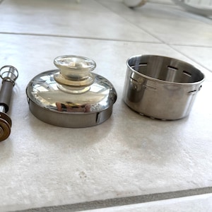 May include: A stainless steel cocktail shaker set, including a lid with a clear glass knob, a cylindrical strainer, and a muddler. The set is designed for mixing drinks and cocktails.