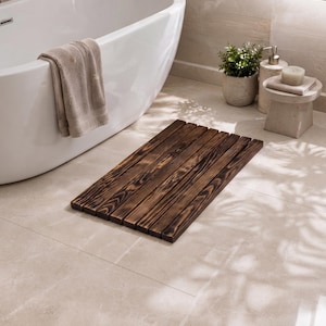 May include: A rectangular bath mat constructed from parallel dark brown wooden slats with a charred, scorched finish. The mat features a series of narrow gaps between each individual plank, creating a slatted surface design for the bathroom floor.
