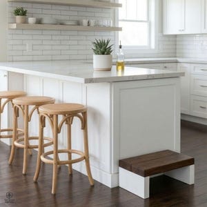 Puede incluir: Una isla de cocina moderna con encimera blanca y gabinetes blancos. Tres taburetes de bar de madera clara con asientos tejidos están frente a la isla. Un pequeño taburete de madera está al lado de la isla. Una planta en maceta y una botella de aceite están en la encimera.