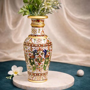 May include: A white marble vase with gold accents and intricate floral and peacock designs. The vase has a slender neck and a wide base, and is filled with green and white faux flowers. The vase is displayed on a round marble stand.