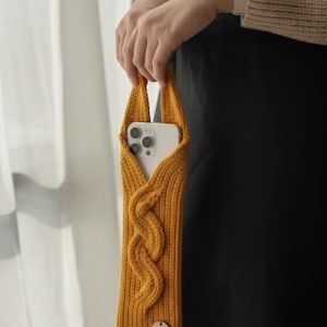 Pode incluir: Capa de telefone de malha amarelo mostarda com design de cabo trançado. A capa tem uma alça de laço e uma pequena etiqueta de madeira. Um smartphone branco é visível dentro.