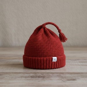 May include: A knitted red beanie with a ribbed brim and a tassel detail. The hat has a rounded crown and a small white tag on the brim. The hat is made of a soft, textured yarn.