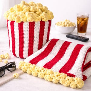 Puede incluir: Un juego de cubo y manta de palomitas de maíz de ganchillo con rayas rojas y blancas. El cubo está lleno de palomitas de maíz amarillas. También se ven unas gafas 3D y un mando a distancia, lo que sugiere un tema de cine.