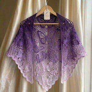May include: A handmade, purple crochet shawl with a butterfly design. The shawl features a gradient of colors, transitioning from deep purple to a light lavender. The intricate openwork design is displayed on a wooden hanger.