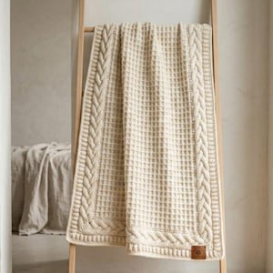 Könnte beinhalten: Eine cremefarbene Strickdecke mit einem zentralen Waffelmuster, das von einem dicken Zopfmuster umrandet ist. Die rechteckige Decke hängt über einer Holzleiter und hat ein kleines braunes quadratisches Etikett an der unteren Ecke.