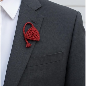 May include: A dark gray suit jacket with a white collared shirt. A handmade, red crocheted lapel flower is pinned to the jacket. The flower has a triangular shape with a tassel and a looped stem.