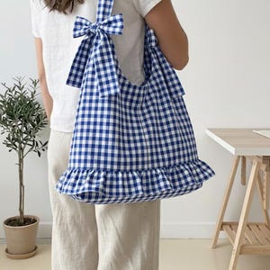 Puede incluir: Bolso tote a cuadros azules y blancos con volantes en la parte inferior y un lazo en la correa. El bolso se lleva al hombro. Un accesorio elegante para el uso diario.