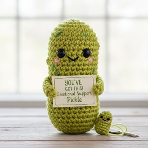 Könnte beinhalten: Ein handgehäkelter, grüner Krokodilgurke mit einem lächelnden Gesicht, das ein Schild mit der Aufschrift "YOU'VE GOT THIS! Emotional Support Pickle" hält. Daneben befindet sich eine kleinere Krokodilgurke. Das Bild enthält auch ein Banner mit der Aufschrift "NEW UNIQUE PATTERN".