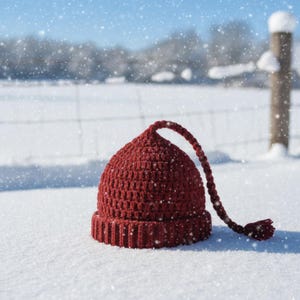 K&ouml;nnte beinhalten: Eine bordeauxfarbene H&auml;kelm&uuml;tze mit langem geflochtenem Kordel und Quaste, die im frischen Schnee liegt. Die M&uuml;tze hat einen gerippten Rand und eine runde Krone. Schneeflocken fallen sanft im Hintergrund, was eine Winterszene andeutet.
