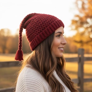May include: A burgundy crochet hat with a long tassel. The hat is a deep red color and has a textured pattern. The tassel hangs down from the side of the hat. The hat is a winter accessory.