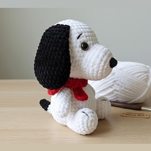 Può includere: Un peluche di Snoopy all'uncinetto fatto a mano. Il cane ha un corpo bianco, orecchie nere e una sciarpa rossa. Il giocattolo è seduto su una superficie di legno. Un gomitolo di lana bianca e un uncinetto sono sullo sfondo.