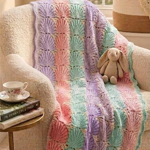 May include: A crochet blanket featuring vertical stripes of shell-patterned stitches in pastel pink, mint green, and lavender. The blanket is draped over a textured cream-colored armchair, with a small plush rabbit sitting on top of the fabric.