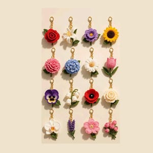 May include: A collection of sixteen individual crochet flower keychains arranged in a grid. Each floral piece features vibrant yarn colours including red, white, purple, yellow, pink, and blue, attached to a gold-toned metal lobster clasp hook.