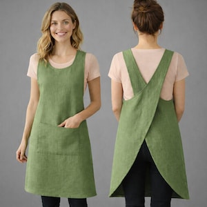 以下が含まれることがあります： グリーンのリネンエプロン。背中はクロスデザインで、大きなフロントポケットと丸いネックラインが特徴です。モデルは薄いピンクのTシャツを着ています。エプロンは膝丈です。