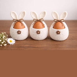 May include: Three white crochet bunny egg holders with brown eggs inside. Each holder has bunny ears, a brown bow, and a small flower. The holders are on a wooden surface, with a small bouquet of white flowers.