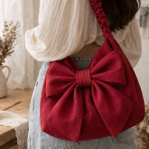 May include: A red fabric shoulder bag with a large bow detail. The bag has a matching red strap and is worn over the shoulder. The bag is made of a textured fabric and is a solid red color.