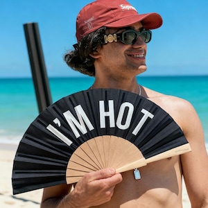 Könnte beinhalten: Ein schwarzer Handfächer mit dem Text "I'M HOT" in Weiß. Der Fächer hat einen Holzgriff und einen kleinen silbernen Anhänger. Der Fächer ist geöffnet und zeigt ein plissiertes Design. Der Hintergrund ist eine Strandszene mit blauem Wasser und Himmel.