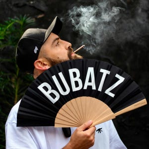 Puede incluir: Abanico de mano negro con la palabra "BUBATZ" en letras blancas grandes. El abanico tiene un mango de madera natural y se sostiene frente a una persona. La persona lleva una gorra negra y una camiseta blanca.