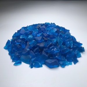 Puede incluir: Un montón de piezas de vidrio azul translúcido de forma irregular. Los fragmentos de vidrio varían en tamaño y reflejan la luz, creando reflejos y sombras. Las piezas están dispersas sobre una superficie blanca.