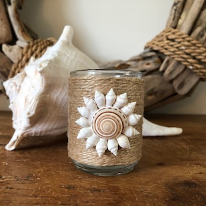 May include: A glass candle holder wrapped in natural twine with a decorative seashell sun design. The holder is on a wooden surface with a large seashell in the background.