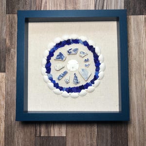 May include: A framed artwork featuring a circular design of white seashells, blue sea glass, and ceramic shards. The arrangement is set against a beige fabric background within a square, navy blue wooden frame. The artwork evokes a coastal theme.