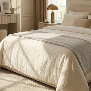 Op de afbeelding: Een crèmekleurige sprei van gestructureerde, linnenachtige stof met een brede, horizontale loper aan het voeteneinde van het bed. De loper heeft een herhalend geometrisch ruitpatroon in lichtgrijze en witte tinten.