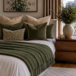May include: A bed featuring a white duvet and a ribbed olive green throw blanket with fringe edges. The bed is layered with several decorative pillows in shades of olive green and cream with floral and geometric patterns, set against a headboard.