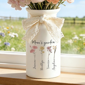 Puede incluir: Un jarrón de cerámica blanco con un lazo de encaje, lleno de flores rosas y melocotón. El jarrón está impreso con las palabras "Mom's garden" e ilustraciones florales, junto con los nombres Jasmine, Brooklyn, Vanessa y Angelina. El jarrón está sobre un alféizar de madera.