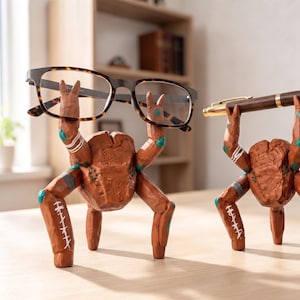 Könnte beinhalten: Zwei braune, geschnitzte, mehrbeinige Figuren mit texturierten Oberflächen, türkisfarbenen Akzenten und weißen Markierungen. Eine Figur hält eine Brille mit Schildpattmuster, die andere einen Kugelschreiber aus dunklem Holz und Gold.