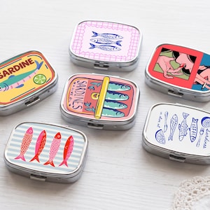 Puede incluir: Seis pastilleros rectangulares de metal con varios diseños temáticos de sardinas. Las tapas muestran ilustraciones coloridas de peces, una rodaja de limón, una persona comiendo y el texto SARDINE, con patrones de rayas o cuadrículas.