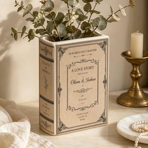 May include: A cream-coloured ceramic vase shaped like a vintage book with black text and ornate borders. The front reads A LOVE STORY WRITTEN BY Olivia & Joshua EST. 08.08.2020. The spine features the names Olivia & Joshua and the text THE BEGINNING OF FOREVER.