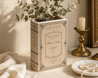 May include: A cream-coloured ceramic vase shaped like a vintage book with black text and ornate borders. The front reads A LOVE STORY WRITTEN BY Olivia & Joshua EST. 08.08.2020. The spine features the names Olivia & Joshua and the text THE BEGINNING OF FOREVER.
