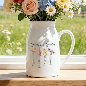 May include: A white ceramic pitcher with a handle, filled with colorful flowers. The pitcher is inscribed with "Grandma's Garden" and floral illustrations with names. The pitcher sits on a wooden surface near a window.